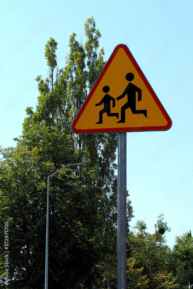 the road sign is made of metal triangular shape in orange color with the image of children against a background of green trees and blue sky . Careful children