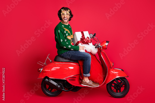 Profile side view portrait of attractive glad cheerful guy riding moped delivering gifts sack St Nicholas tradition shop order service isolated bright vivid shine vibrant red color background