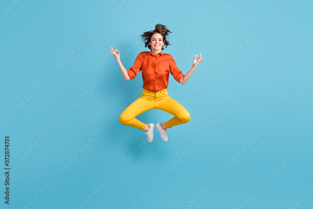 Full length photo of attractive funny lady jump high up positive energy emotions practice yoga om position in flight wear orange shirt yellow trousers sneakers isolated blue color background