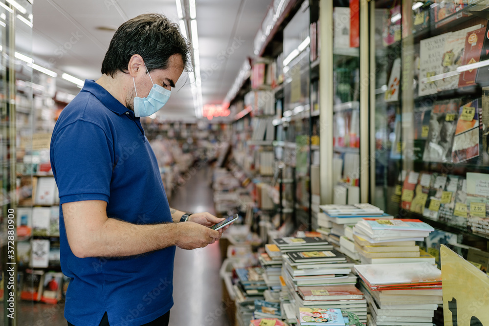 Middle-aged adult wearing hygiene face mask checking the smartphone in a bookstore next to second-hand books. Purchase of a back-to-school textbook. Parent