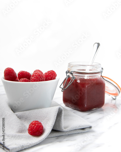 Raspberry jam in a glass jar with spoon and a cup of ripe raspberries with tablecloth on marble table. Front view food photography