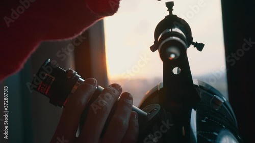 Girl next to the window looking through the telescope at the sky in her bedroom at sunset. Hobbie while you have to be locked up at home. Confinement, being at home.