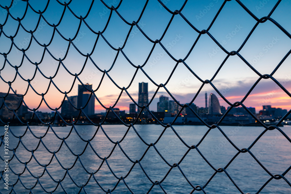 Fototapeta premium Cityscape by the sea through the grid of fence during sunset. Selective focus.