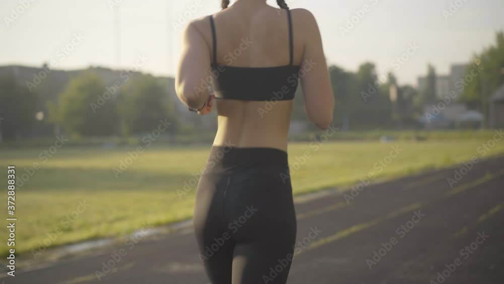 Camera follows young Caucasian jogger training in sunlight outdoors. Confident woman launching app on smart watch and start running along the stadium. Morning training of slim runner at sunrise.