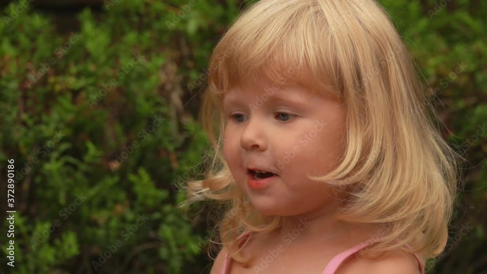 Beautiful little blond girl is smiling and closing her mouth with her hand in amazement in the sunny park outdoors Beautiful little blond girl is smiling and closing her mouth with her hand in amazement in the sunny park outdoors
