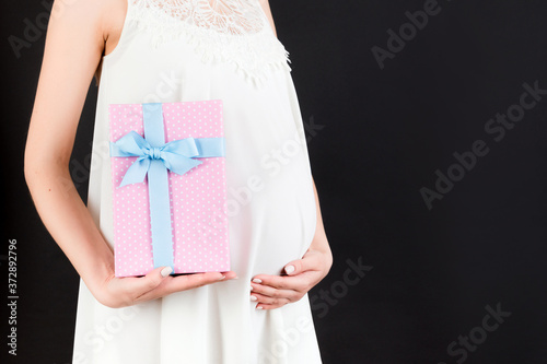 Close up of pregnant woman in white dress holding pink spotted gift box at black background. Expecting a baby girl. Copy space © sosiukin