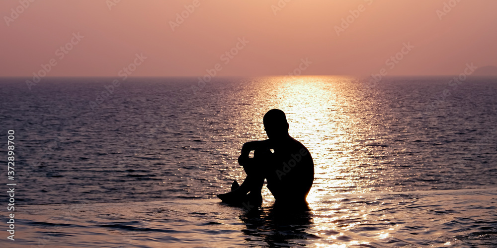Young boy is sitting in a pool enjoying the sunset. Human silhouette over the setting sun. Swimming pool in the evening.