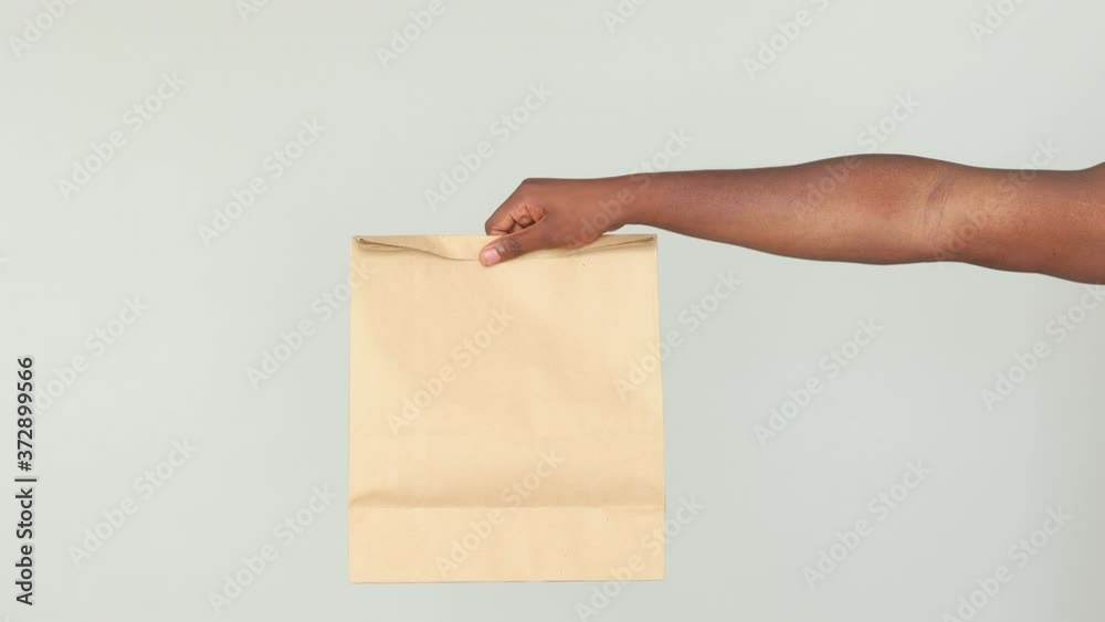 Close up african man male guy hand hold brown blank craft paper bag takeaway mock up copy space isolated on gray background. Food products delivery courier service from shop restaurant to work office Close up african man male guy hand hold brown blank craft paper bag takeaway mock up copy space isolated on gray background. Food products delivery courier service from shop restaurant to work office