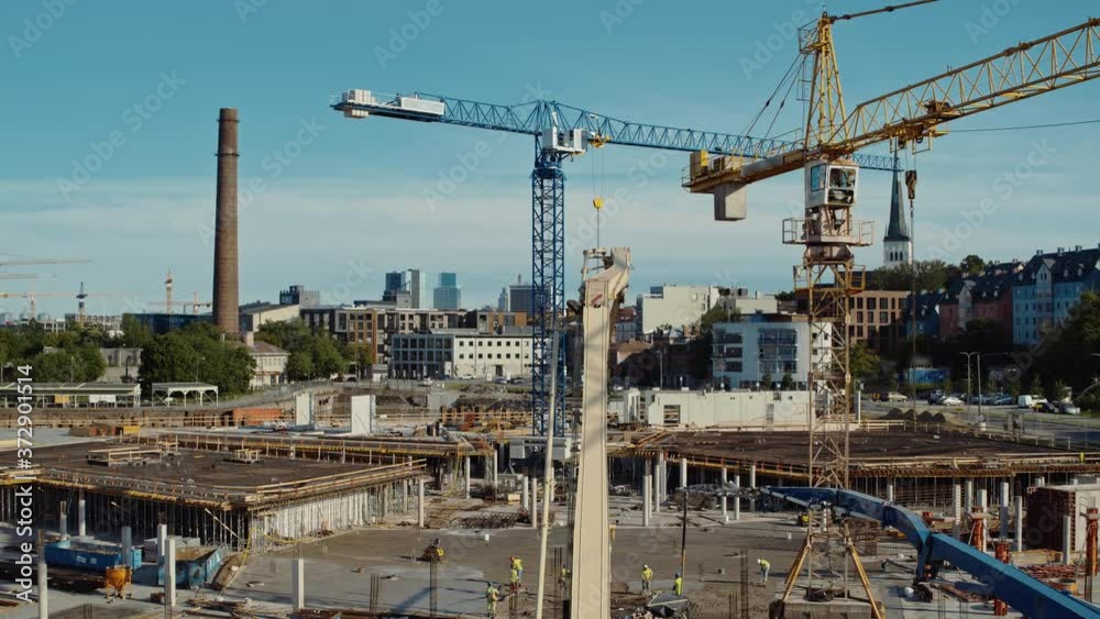 Aerial Flight Over a New Constructions Development Site with High Tower Cranes Building Real Estate. Heavy Machinery and Construction Workers are Employed. City Houses in the Background. Aerial Flight Over a New Constructions Development Site with High Tower Cranes Building Real Estate. Heavy Machinery and Construction Workers are Employed. City Houses in the Background.