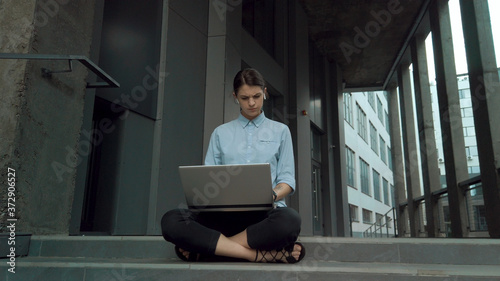 Business woman with laptop sitting on stairs of business center. Female office worker typing text on laptop