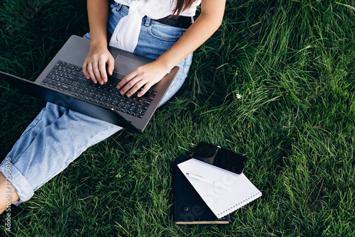 student girl with laptop outdoors sitting on the grass, surfing the internet or preparing for exams. Technology, education and remote work concept. Soft selective focus.