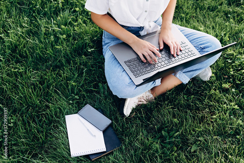 student girl with laptop outdoors sitting on the grass, surfing the internet or preparing for exams. Technology, education and remote work concept. Soft selective focus.