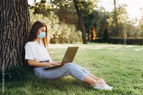 beautiful and young girl who studies and works with a laptop with a surgical mask in the park, blogger and smart worker during the epidemic, home schooling, distance learning. Soft selective focus. © Tasha Sinchuk