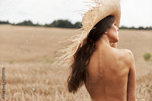 photo of the back of a beautiful tanned girl who has a straw hat on head, dark hair in ponytail, she looks away standing on a wheat field