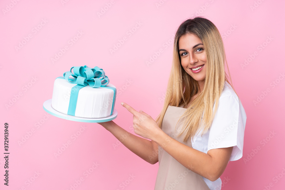 Young Uruguayan pastry woman with a big cake over isolated pink background and pointing it