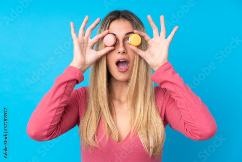 Young Uruguayan woman over isolated blue background holding colorful French macarons with surprised expression