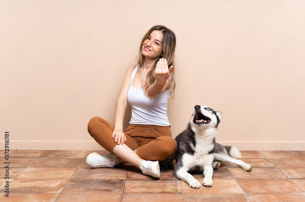 Young pretty woman with her husky dog sitting in the floor at indoors inviting to come with hand. Happy that you came