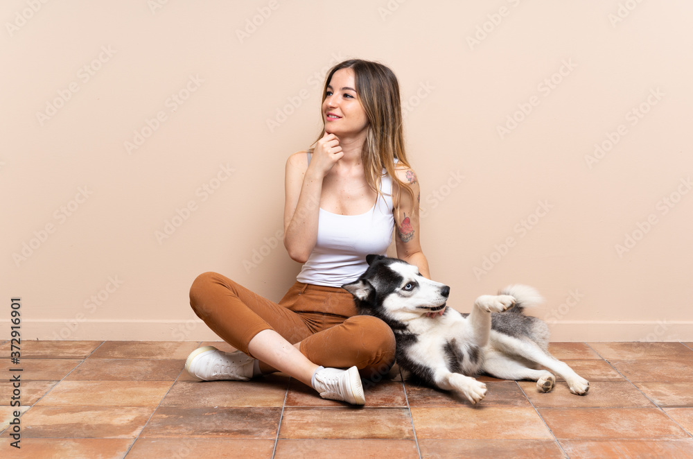 Young pretty woman with her husky dog sitting in the floor at indoors looking to the side and smiling Young pretty woman with her husky dog sitting in the floor at indoors looking to the side and smiling