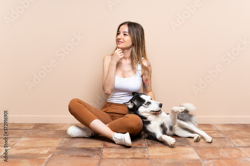Young pretty woman with her husky dog sitting in the floor at indoors looking to the side and smiling