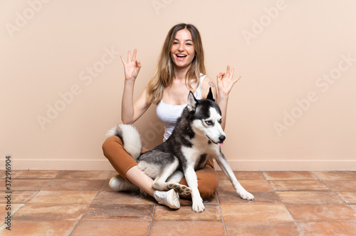 Young pretty woman with her husky dog sitting in the floor at indoors showing ok sign with two hands