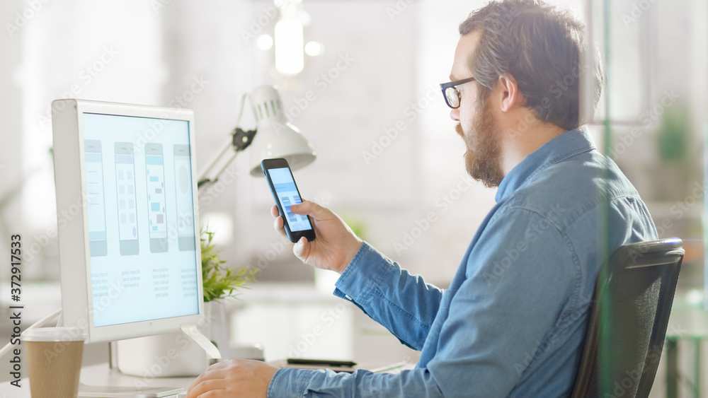 Young Focused Software Developer Compares Smartphone Application Design with Same App Design Shown on Desktop Computer Screen. He is Working in a Bright Modern Loft Office. Young Focused Software Developer Compares Smartphone Application Design with Same App Design Shown on Desktop Computer Screen. He is Working in a Bright Modern Loft Office.