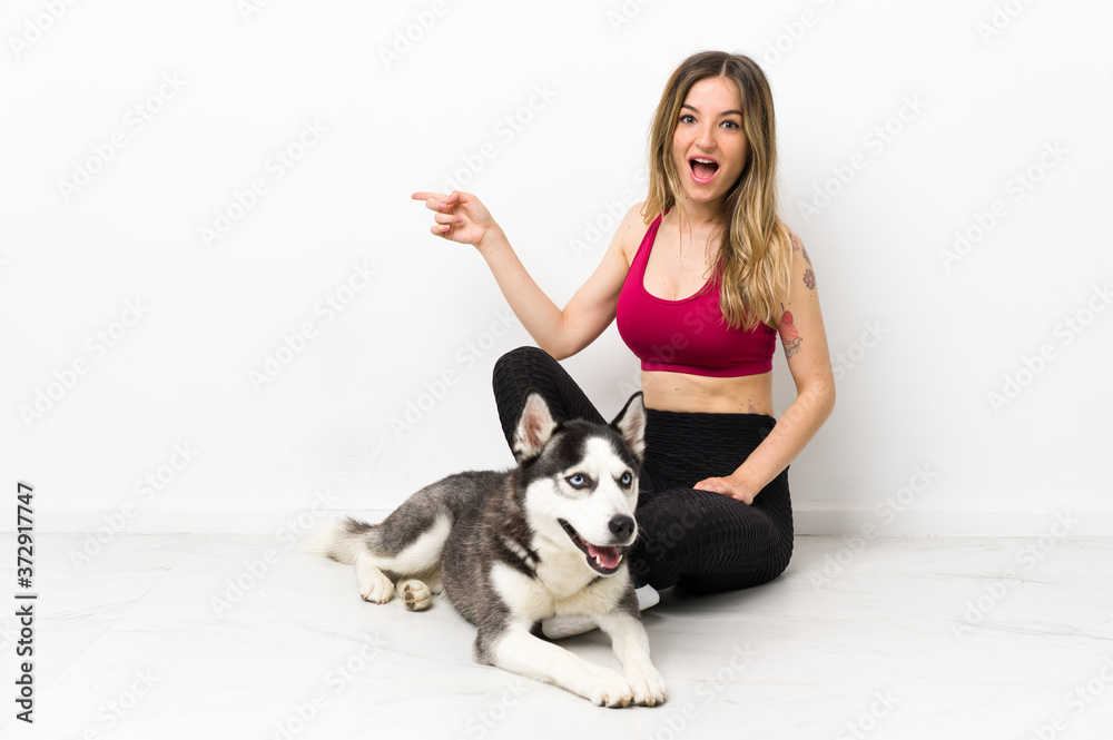 Young sport girl with her dog sitting on the floor surprised and pointing finger to the side Young sport girl with her dog sitting on the floor surprised and pointing finger to the side