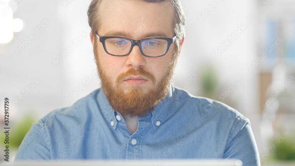 Fototapeta premium Portrait shot of a Bearded Creative Young Man Wearing Glasses Sitting in His Bright Office and Working on a Personal Computer. Computer Screen Reflects in His Glasses.