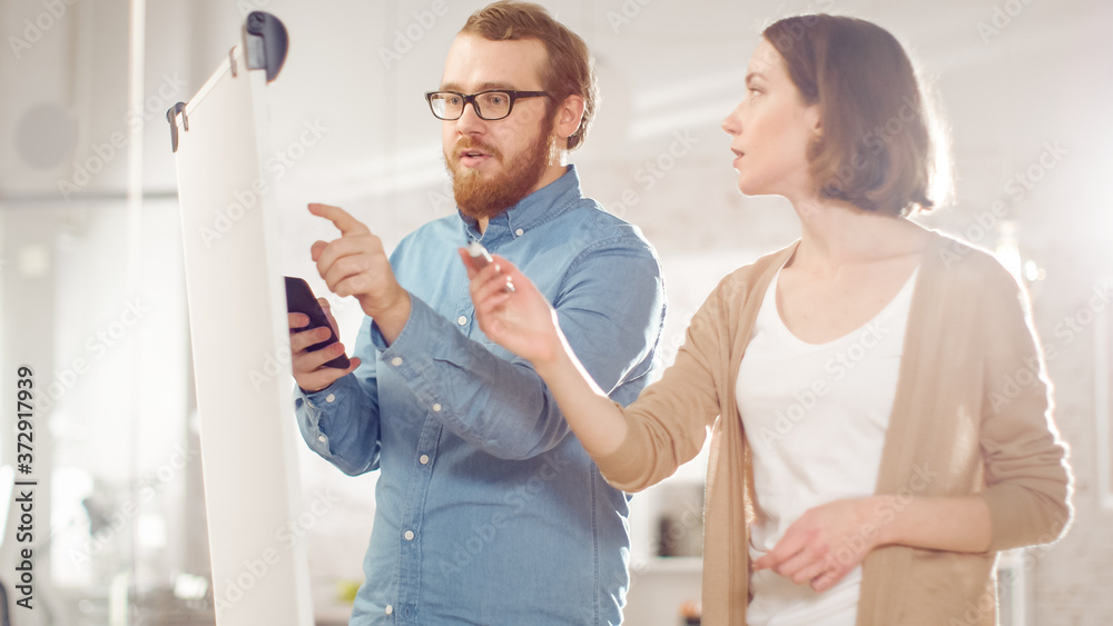 Obraz premium Young Man and a Woman Discuss Working Process on a Whiteboard. Man Holds Smartphone, woman Draws on a Whiteboard. Bright Modern High Tech Office with Beautiful People.