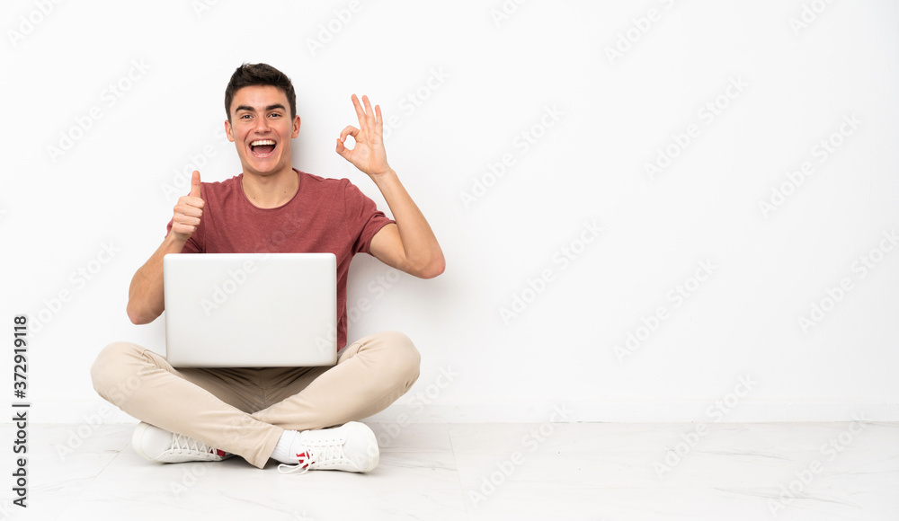 Teenager man sitting on the flor with his laptop showing ok sign and thumb up gesture Teenager man sitting on the flor with his laptop showing ok sign and thumb up gesture