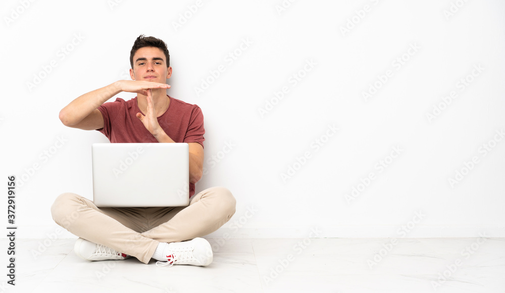 Teenager man sitting on the flor with his laptop making time out gesture Teenager man sitting on the flor with his laptop making time out gesture