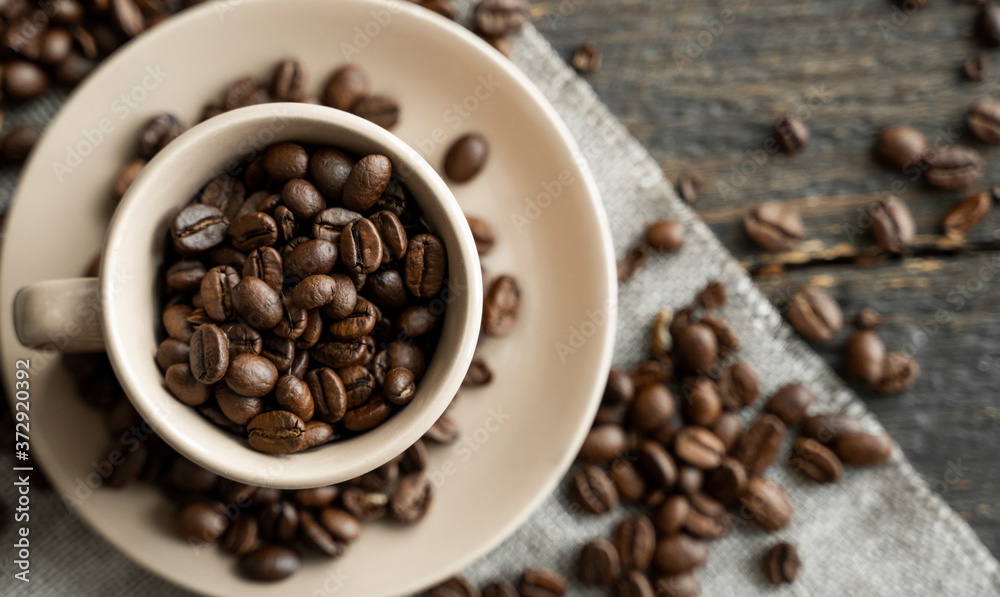 Coffee cup filled of fresh arabica or robusta coffee beans with scattered coffee beans on a linen textile and wood table. Coffee cup filled of fresh arabica or robusta coffee beans with scattered coffee beans on a linen textile and wood table.