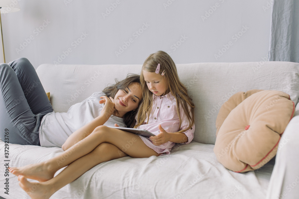 Mother and little daughter watching cartoons together on cozy sofa at home. 