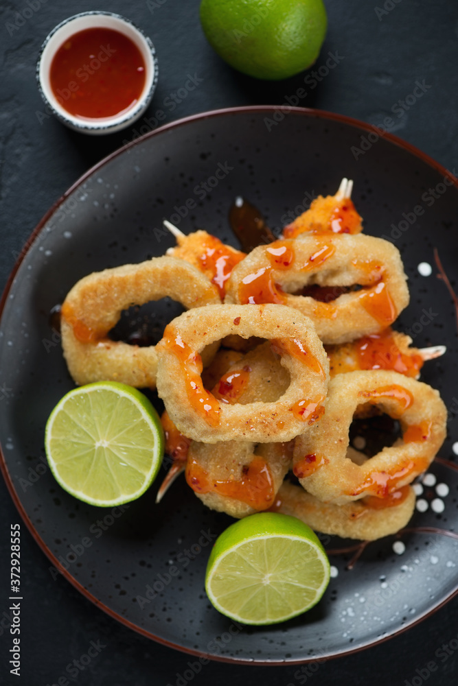 Black plate with fried panko breaded calamari rings and crab claws, lime and dipping sauce, vertical shot, top view