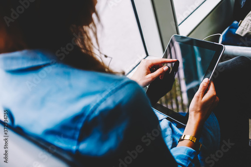 Back cropped view of young woman touching with finger on display of modern touch pad chatting online via public transport wifi.Female person holding tablet with earphones in hands sitting in tram © BullRun