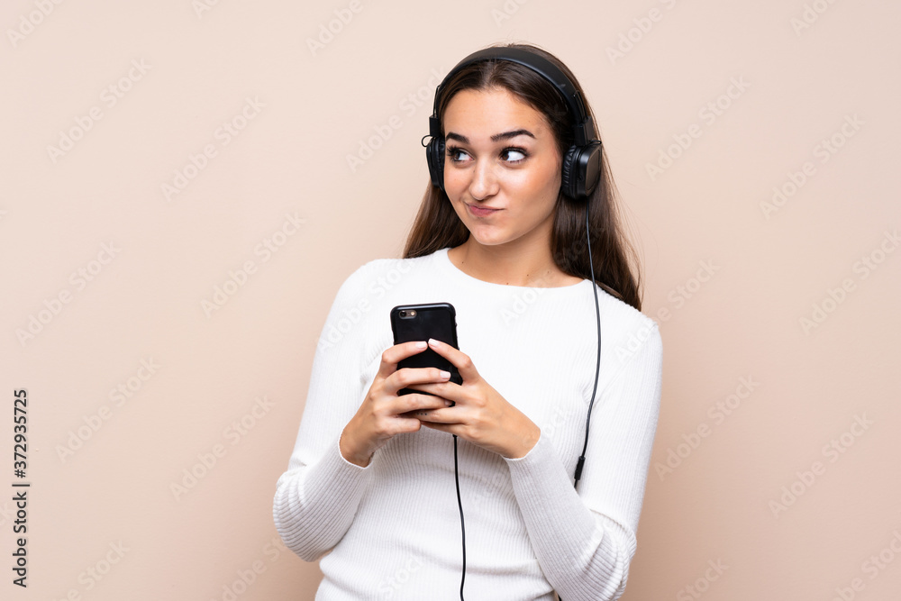 Young woman over isolated background using the mobile with headphones and thinking Young woman over isolated background using the mobile with headphones and thinking