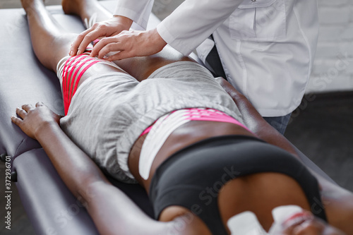 Kinesiology taping. Physiotherapist applying kinesiology tape to patient hips.Therapist treating young female African American athlete. Post traumatic rehabilitation, fat lose,sport physical therapy.