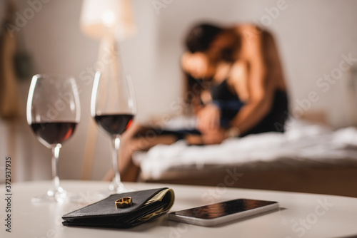 Selective focus of wedding ring on wallet near smartphone and glasses of wine with shirtless man embracing woman on bed photo
