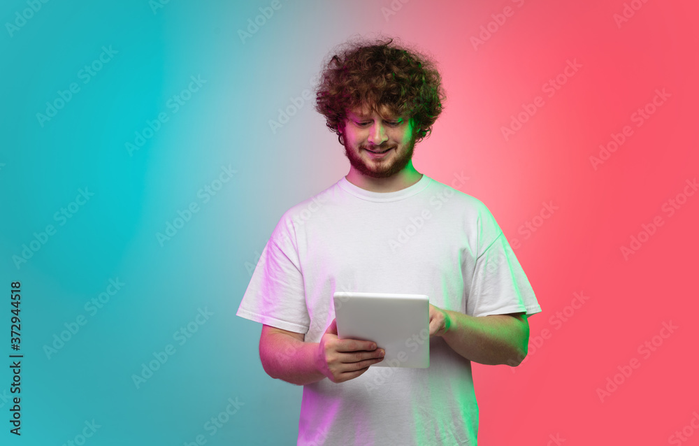 Scrolling tablet. Caucasian young man's portrait isolated on gradient studio background in neon light. Beautiful male model in casual style. Concept of human emotions, facial expression, youth, sales