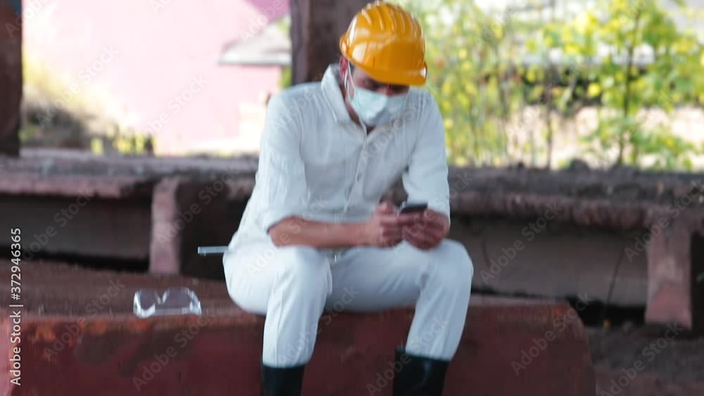 A male engineer wearing a mask on his face uses the phone during a break from work. View news and social networks. Relaxation of the employee's psyche. A male engineer wearing a mask on his face uses the phone during a break from work. View news and social networks. Relaxation of the employee's psyche.