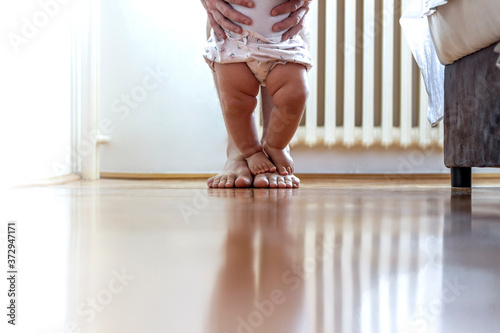 Baby girl walking on wooden floor. First steps of a child with her father's help. Both barefoot on hardwood floor. Cropped image a father teaching baby girl how to walk.Girl taking her first steps. photo