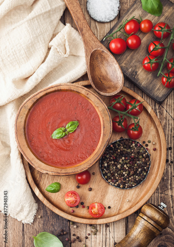 Wooden plate of creamy tomato soup 0n round tray, pepper and kitchen cloth on wooden background with box of raw tomatoes.