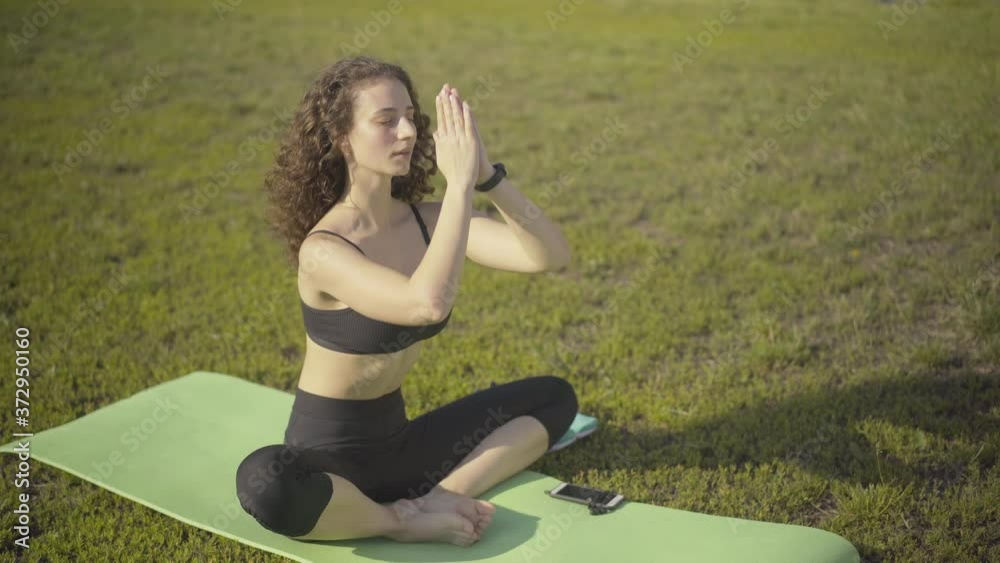 Relaxed curly-haired woman putting hands together and breathing out. Portrait of confident Caucasian yogi training on exercise mat in summer morning outdoors. Meditation and workout concept. Relaxed curly-haired woman putting hands together and breathing out. Portrait of confident Caucasian yogi training on exercise mat in summer morning outdoors. Meditation and workout concept.