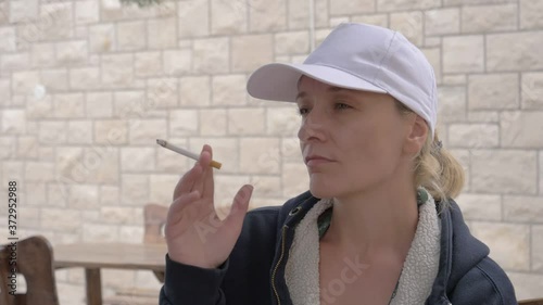 A young woman in a cap takes a cigarette smoke and blows it out through her mouth. Slow motion 120fps converted to 24 fps. Smoking a cigarette concept.
