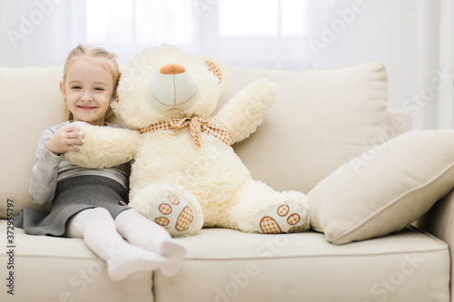 Portrait of a young smiling girl with stuffed toy sitting on sofa in the living room at home.