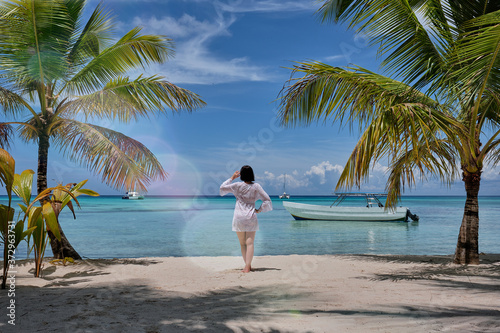 A girl looking horizon standing next to a palm trees on a beach in front of a Caribbean sea