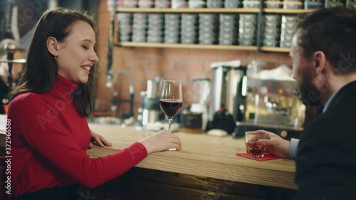 Beatiful yound woman in red clothes sitting next to bar counter in hookah lounge and talking with man in business suit laughing and holding glass of red wine.