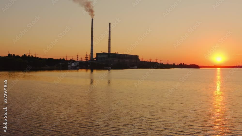 Evening river on power plant background. Smoke from industrial pipes goes into the air against the setting orange sun. Silhouette of a factory near the lake. Evening river on power plant background. Smoke from industrial pipes goes into the air against the setting orange sun. Silhouette of a factory near the lake.