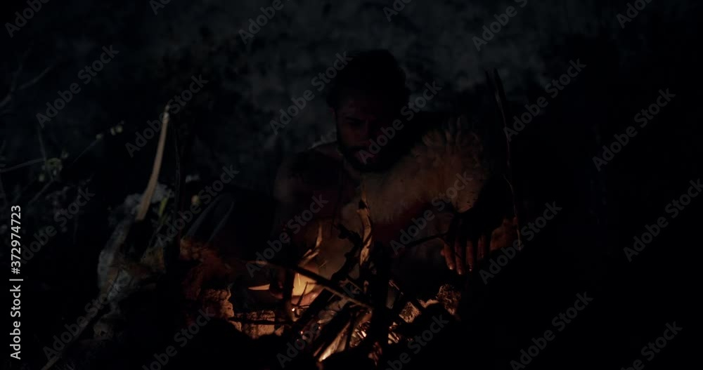 Bearded man in animal hide adjusting firewood and putting apple in campfire while resting and cooking at dark night in wilderness. Reproduction of the historical life of Neanderthal man, or Cro-Magnon