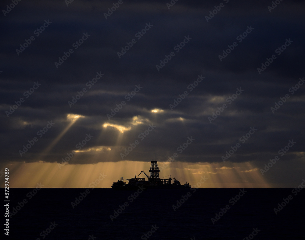 Backlit oil rig at dawn, cloudy sky and beams of light through the clouds