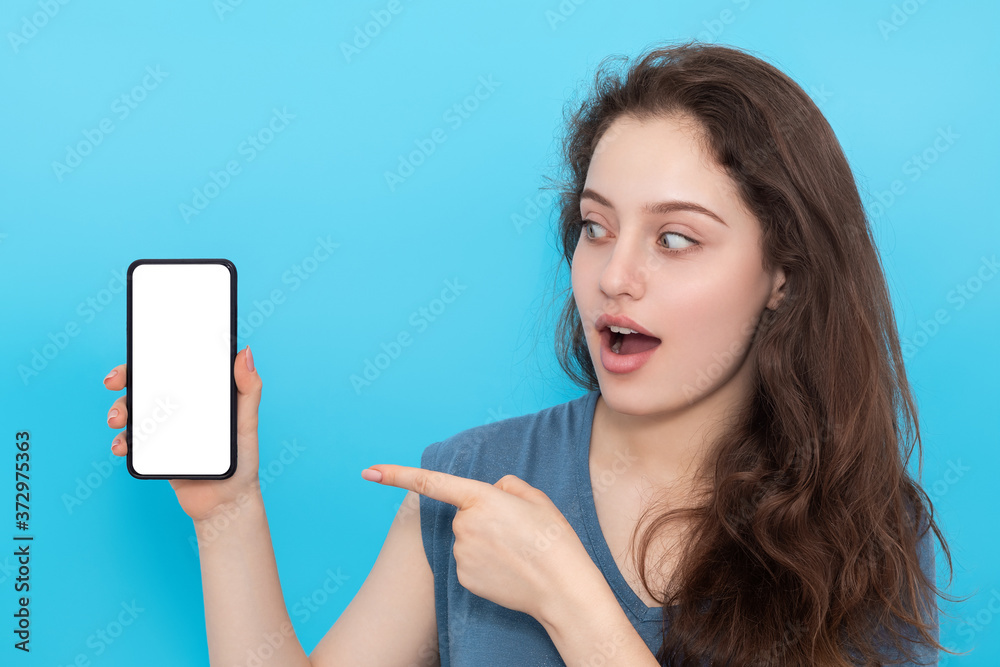 Young woman with surprise shows a finger to a smartphone in her hand. Young girl with long hair and blue eyes. Young woman with surprise shows a finger to a smartphone in her hand. Young girl with long hair and blue eyes.